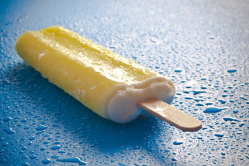 lemon ice lolly lying on blue background with ice and drops of water