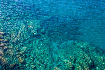 From the Cinque terre trail aerial view of the transparent, emerald, blue sea and the young woman floating in it.