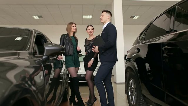Two gorgeous girls talking to a sales consultant in a car dealership, they want to buy a new car. The process of working in the showroom.