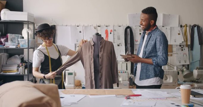 Female tailor is measuring garment while African American man her colleague is writing in notebook talking smiling. Fashion design and handmade clothes concept.