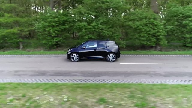 Aerial side view footage of black electric vehicle or EV in short driving over straight road with trees on the side showing sunshine reflection on the car very stable and precise point of view 4k