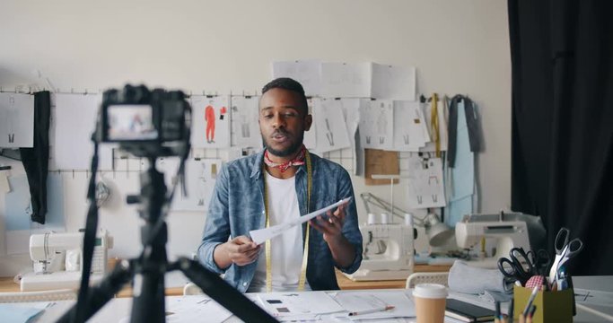 Male fashion designer vlogger recording video for internet blog showing sketches speaking and gesturing working in studio alone. People and vlogging concept.