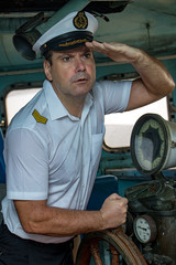 A sailor officer in the uniform is steering the ship with a rudder. Captain standing in the wheelhouse of ship and watching the surroundings. © milkovasa
