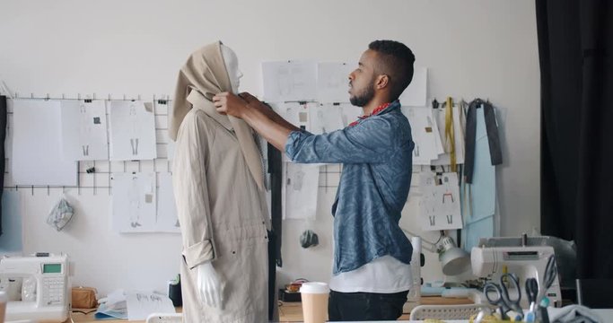 Male African American designer is dressing mannequin then looking and smiling enjoying stylish garment working alone in workshop. Creative people and fashion concept.