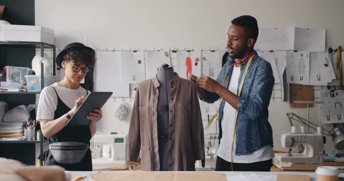 Tailor African American man is measuring clothes on mannequin while Caucasian girl colleague working with tablet assisting. Fashio design and people concept.