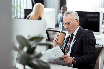 selective focus of handsome businessman reading newspaper while holding disposable cup near multicultural coworkers