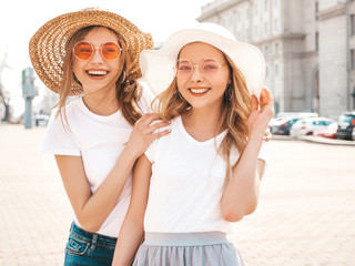 Portrait of two young beautiful blond smiling hipster girls in trendy summer white t-shirt clothes. Sexy carefree women posing on street background. Positive models having fun in sunglasses and hat