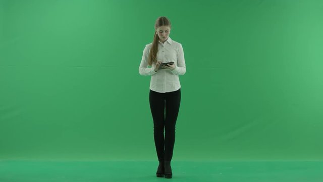Young business woman is chating on her tablet. She wears formal dress: white shirt and blak trousers and stands on the green screen.