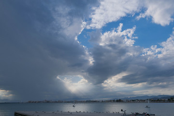 Dark heavy clouds covered the blue sky over Mediterranean sea. Hard rain clouds. Sun beam through heavy sky, gulls sit, sailing boats go to the port.