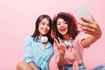 Pleased tanned asian girl gently smiling while her african friend making selfie. Indoor portrait of glad black woman with smartphone taking picture of herself on pink background near hispanic lady.