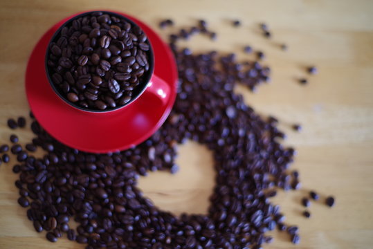 Top view of roasted coffee beans in red cup with pile of beans lie in the shape of a heart on the wooden table.