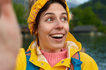 Close up shot of optimistic female with broad smile, looks away, dressed in yellow raincoat, spends time near small pond or river, makes selfie portrait, carries rucksack, spends free time outdoor