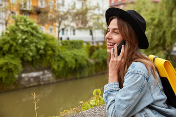 Sideways shot of happy Caucasian has pleasant talk on cell phone, poses near small river in ancient city, carries yellow rucksack on back, wears hat, denim jacket, focused into distance joyfully