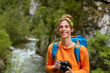 Positive backpacker covers distance, looks far away with glad dreamy expression, holds camera,...