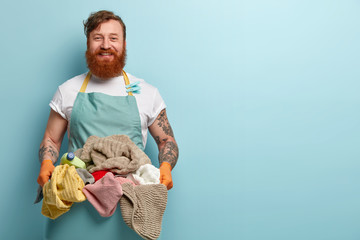Glad satisfied red haired man happy finish domestic work, holds pile of fresh clean laundry, wears casual t shirt with apron and clothespins, protective gloves, stands over blue background, free space