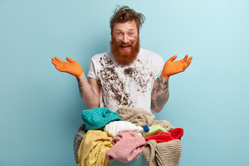 Happy red haired man with thick bristle, spreads hands, has fun after doing laundry at home,...