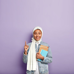 Smiling dark skinned woman has Islamic view, wears white hijab, points with fore finger upwards,...
