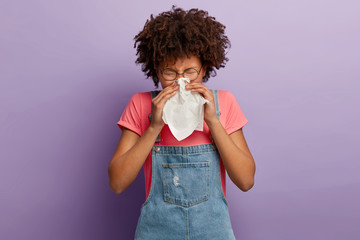 Curly young female feels unwell, blows nose in white tissue, suffers from running nose, cold symptoms or allergy, sneezes very often, wears round spectacles, casual t shirt and denim sarafan - obrazy, fototapety, plakaty