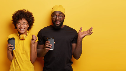 Happy students have coffee break, drink beverage from disposable cup, wear casual clothes, round spectacles, gesture from joy, stand closely to each other against yellow wall, empty space on right