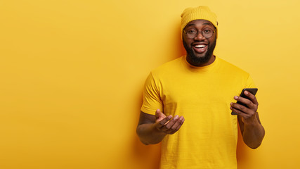 Studio shot of smiling carefree dark skinned man updates profile in social networks, holds modern smartphone device in hand, wears casual t shirt and hat, stands over yellow wall with blank space area