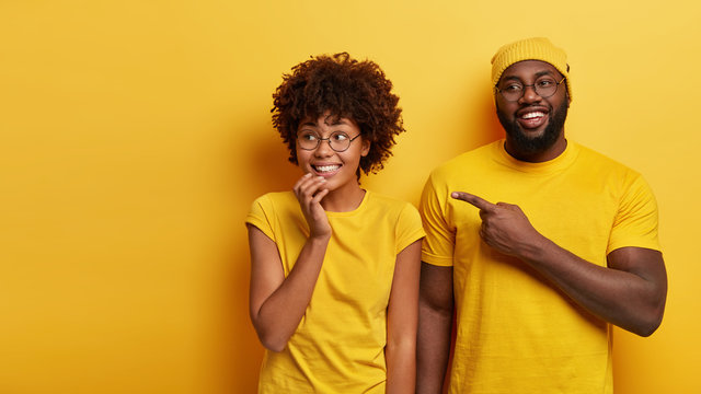 Studio Shot Of Happy Young Woman And Man Look Happily Aside, Point At Blank Space, Notices Awesome Product, Stand Against Yellow Studio Background. People, Advertisement And Ethnicity Concept
