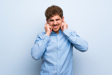 Blonde man over blue wall frustrated and covering ears