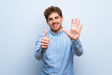 Blonde man over blue wall counting six with fingers