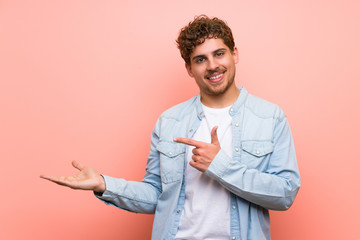 Blonde man over pink wall holding copyspace imaginary on the palm to insert an ad