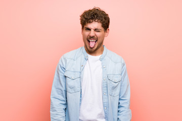 Blonde man over pink wall showing tongue at the camera having funny look