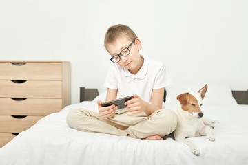 boy with a dog jack russell playing on a smartphone on a white bed at home