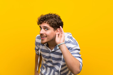 Blonde man over yellow wall listening to something by putting hand on the ear
