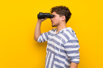 Blonde man over yellow wall and looking in the distance with binoculars