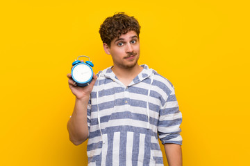 Blonde man over yellow wall holding vintage alarm clock