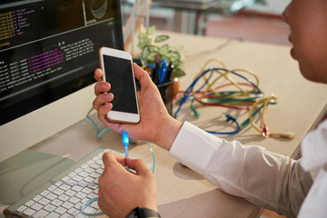 Close-up of young programmer sitting at the table in front of computer monitor with software and connecting his mobile phone with usb cable