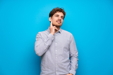 Blonde man over blue wall thinking an idea while looking up