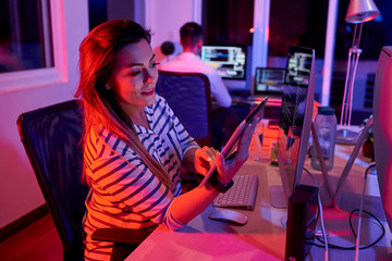 Smiling young businesswoman sitting at workplace in front of computer monitor and working online using digital tablet in dark office with businessman working in the background.