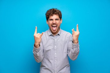 Blonde man over blue wall making rock gesture