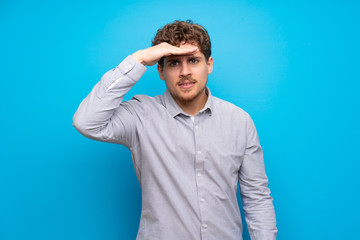 Blonde man over blue wall looking far away with hand to look something