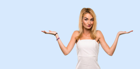 Young blonde woman having doubts while raising hands over isolated blue background