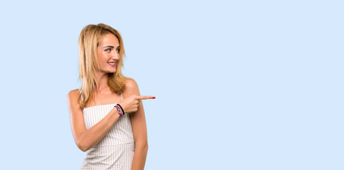 Young blonde woman pointing finger to the side over isolated blue background