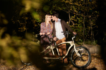 Happy romantic couple, bearded man and attractive girl close together at tandem double bicycle outdoors in dark autumn park or forest on sunny clearing in the fall