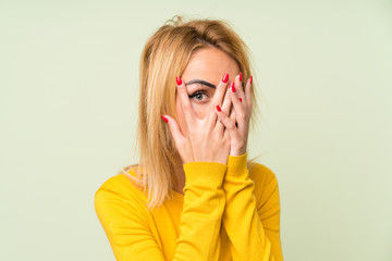 Young blonde woman over isolated green background covering eyes and looking through fingers