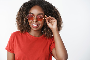 Girl ready for vacation and you. Portrait of charming carefree enthusiastic african-american female with curly haircut wearing stylish red sunglasses and gazing upbeat at camera over white wall