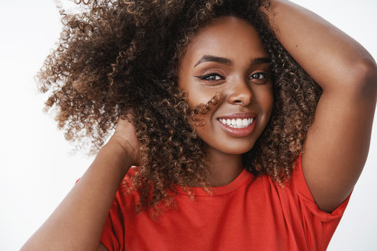 Close-up shot of carefree relaxed and tender african-american female model in red t-shirt playing with curly hair holding hands on head tilting and smiling caring with sensual gaze over white wall