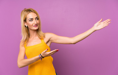 Young blonde woman over purple background extending hands to the side for inviting to come