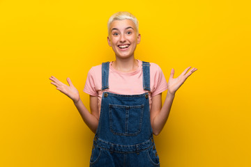 Teenager girl with overalls on yellow background with shocked facial expression