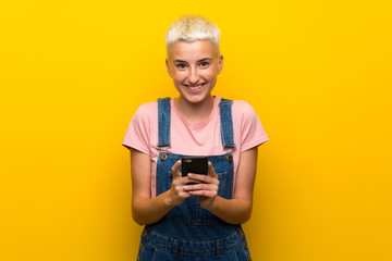 Teenager girl with overalls on yellow background sending a message with the mobile