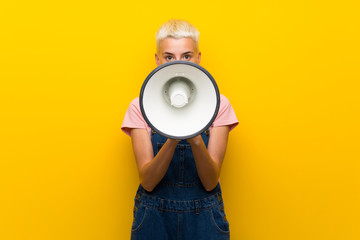 Teenager girl with overalls on yellow background shouting through a megaphone