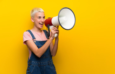 Teenager girl with overalls on yellow background shouting through a megaphone