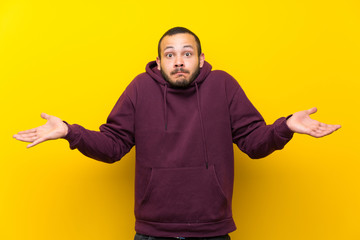 Colombian man with sweatshirt over yellow wall having doubts while raising hands